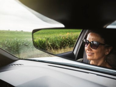Gülümseyen bir kadının yüzü araba sürerken dikiz aynasından yansıyor ve manzaranın tadını çıkarıyor..