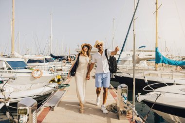 Neşeli bir çift marinada güneşli bir günün tadını çıkarıyor, rıhtımda teknelerle çevrili yürüyor..