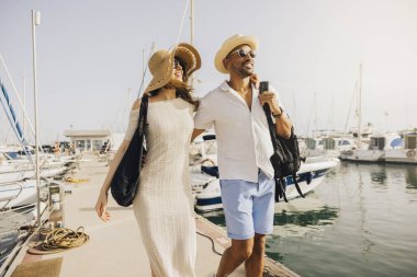 Gülümseyen bir çift, güneşli bir günde marina rıhtımında teknelerle el ele yürür..