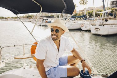 Güneş gözlüğü takan ve şapka takan rahat bir adam teknelerle dolu bir limanda güneşli bir günün tadını çıkarıyor..