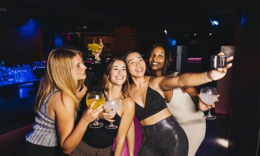 A group of four young women happily take a selfie together while enjoying drinks at a vibrant party. clipart