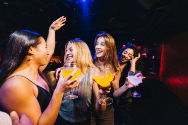 Bir grup gülen arkadaş bir gece kulübünde içki içip gülüyorlar. Atmosfer canlı ve sosyaldir. Havada sevinç ve kutlama duygusu vardır..