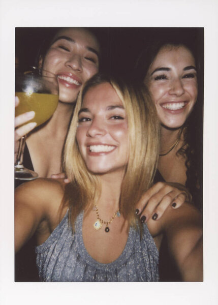 Three smiling friends enjoy a festive night, captured with an instant camera selfie. The image has a vintage look with dusty and scratched effects, highlighting joy and friendship.