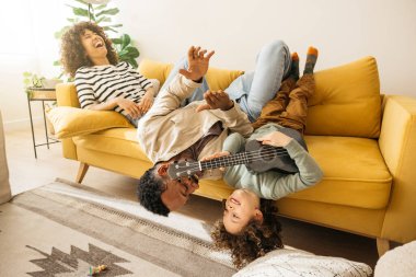 Bir ailenin birlikte çaldığı, ukulele tutan bir çocuğun olduğu neşeli bir an. Sahne sıcaklığı ve mutluluğu resmediyor, rahat bir kanepede vakit geçiriyorlar..