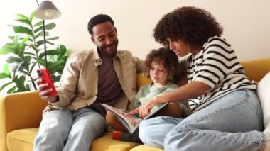 Neşeli bir aile rahat bir kanepede birlikte oturuyor, evde kitap okurken çocuklarıyla meşgul oluyorlar..