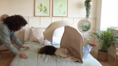 Anne ve oğlu, rahat, modern bir yatak odasında, bir yatağın üzerinde battaniyeyle oynuyorlar..