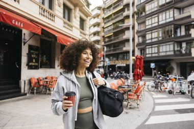 Neşeli bir kadın elinde kahve ve çantayla geziyor, canlı bir şehir kafesinin yanında, spor salonuna gidiyor..