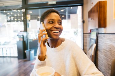Kahve molası veren genç bir kadın, rahat bir kafede akıllı telefonuyla mutlu bir şekilde sohbet ediyor..