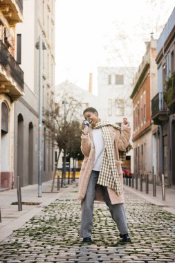 Modaya uygun genç bir kadın kahvesini yudumlar ve büyüleyici bir kaldırım taşı caddesinde dans eder.