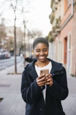 Gülümseyen genç bir kadın sokakta yürürken cep telefonu kullanıyor.