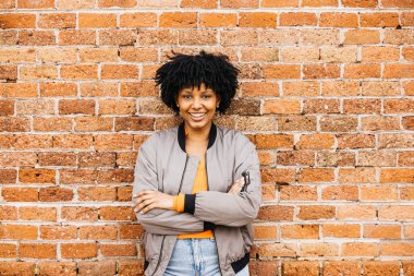 Tuğla bir duvara dayanmış neşeli genç bir kadının portresi. Kollarını kavuşturmuş, kendine güveni ve stili tam.