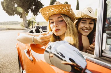 İki arkadaş güneşli bir yaz gününde üstü açık bir arabada seyahat ederken ruj sürüyorlar.