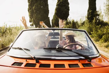 İki turist, yaz tatilinde kırsal alanda, kolları havada, üstü açık, klasik bir araba kullanıyorlar.