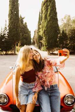 İki arkadaş, yaz gezisi sırasında eski bir arabaya yaslanmış akıllı telefonlarıyla selfie çekiyorlar.