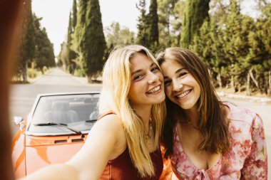 İki mutlu bayan arkadaş yaz gezisi sırasında üstü açık klasik arabalarının yanında selfie çekiyorlar.