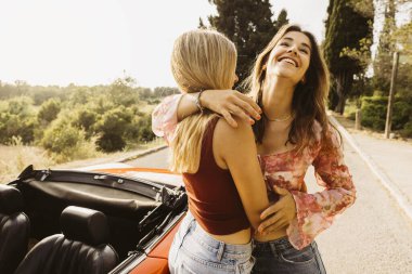 İki bayan arkadaş, eski model üstü açık arabalarıyla bir yaz gezisi sırasında sarılıp gülüyorlar.