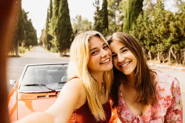 İki mutlu bayan arkadaş yaz gezisi sırasında üstü açık klasik arabalarının yanında selfie çekiyorlar.