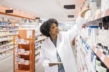 Genç eczacı dijital tablet kullanıyor. Envanteri kontrol ediyor ve modern eczanenin raflarında ürünlere ulaşmaya çalışıyor.