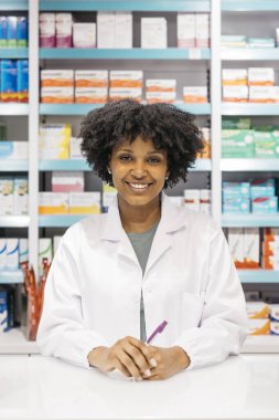 Genç, Afro-Amerikalı Amerikalı eczacı elinde kalemle modern eczanede tezgahta dururken gülümsüyor.