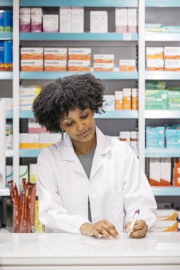 Eczaneye notlar yazan konsantre bir eczacı. Arkaplanda ilaç kutularıyla dolu raflar var.