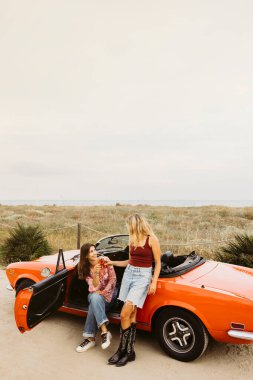 İki arkadaş eski arabalarıyla bir anı paylaşıyor, plaj tatili boyunca içkilerinin tadını çıkarıyorlar.