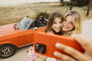 İki arkadaş yaz gezisi sırasında üstü açık bir arabanın önünde akıllı telefonlarıyla selfie çekiyorlar.