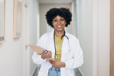 Kendine güvenen doktor, elinde bir not panosu ve kalem tutarken gülümsüyor. Klinik ortamında profesyonellik ve sağlık uzmanlığı gösteriyor.