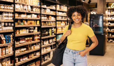 Gurme bir süpermarkette alışveriş yaparken gülümseyen ve tekrar kullanılabilir bir çanta taşıyan genç kadın.