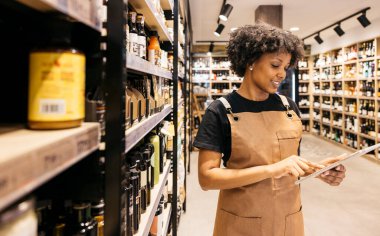 Şarap rafları ve kaliteli gıda ürünleriyle çevrili bir süpermarkette envanter yönetimi için tablet kullanan gülümseyen bir satıcı.