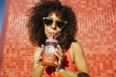 Kıvırcık saçlı, güneş gözlüğü takan bir kadın yaz günü kavanozdaki pipetten ferahlatıcı bir içecek içiyor.