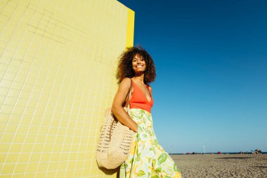 Kumsalda sarı fayanslı duvara yaslanmış, saman torbası ve renkli etekle gülümseyen genç bir kadın.