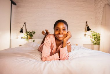 Genç bir kadın pijamalarıyla karnının üzerinde uzanmış, rahat yatak odasında kameraya gülümsüyor.