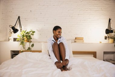 Genç siyahi kadın gülümsüyor, minimalist yatak odasında yatakta rahatça oturuyor.