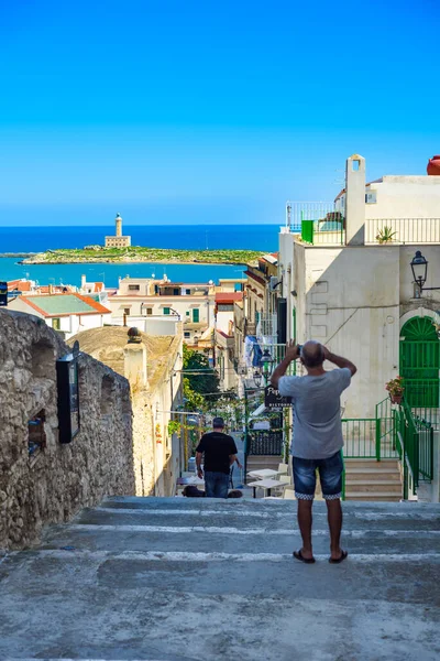 Vieste, İtalya. Via Cesare Battisti 'den kasaba manzarası. Uzakta Vieste 'nin deniz feneri. Bir adam panoramanın fotoğrafını çekiyor. Dikey görüntü. 5 Eylül 2022.