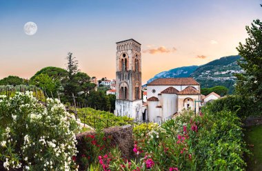 Ravello, İtalya. Santa Maria Assunta Katedrali 'nin üzerinde nefes kesici bir gün batımı ve çiçeklerle renkli zakkum çiçekleri arasındaki çan kulesi. Büyük Ay solda. 2020-08-09.