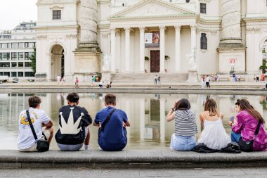 Viyana, Avusturya. St. Charles Kilisesi (Karlskirche) cephesi. Ön planda, bir grup genç arkadaş kilisenin yansıtıldığı çeşmenin kenarında oturuyorlar. 2023-08-02.