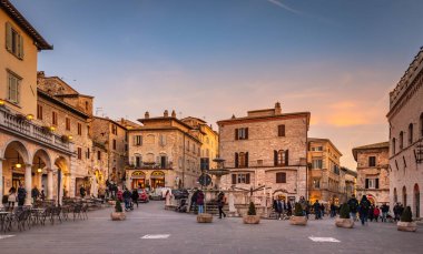 Assisi, İtalya. Akşam üstü Piazza del Comune manzarası. Sokaklarda bir sürü insan ve turist dolaşıyor. Ortada Üç Aslan Çeşmesi var.. 