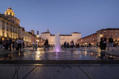 Torino, İtalya. Piazza Castello 'da su sıçrayan ve renkli su oyunları. Kraliyet Müzeleri 'nin ve diğer tarihi binaların ana cephesi gece aydınlandı. Meydanda toplanan insanlar. 2024-11-02.