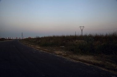 Gün batımında bir köy yolunun kenarındaki toprak setinin arkasındaki alanda elektrik direği.