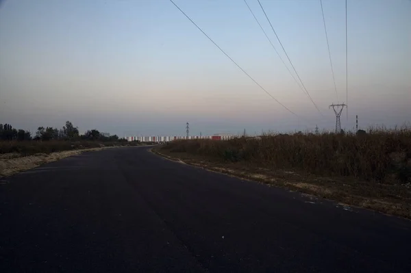 日落时乡间道路边的堤岸后面的田里的电塔 — 图库照片