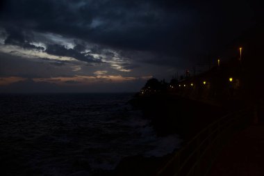 Alacakaranlıkta deniz kenarındaki bir uçurumda sokak ışıklarıyla panoramik yürüyüş