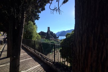 Malcesine 'deki Scaligero şatosunun kulesi ağaçlar ve zakkum bitkileriyle çevrili ve arka planda bir dağ sırtı var.