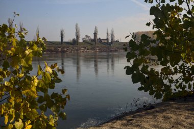 İtalya 'nın kırsal kesimindeki bitki ve çalıların çerçevelediği bir barajın arkasındaki nehir kenarındaki çan kulesi.