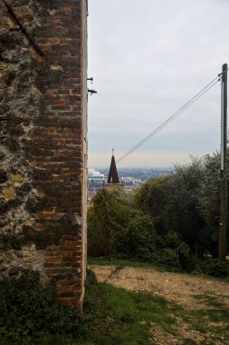 Bulutlu bir günde zeytin ağaçlarının çerçevelediği uzaklıktaki çan kulesi ve antik bir duvarın yanındaki bir tepedeki toprak yol.