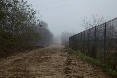 Sisli bir günde metalik bir çitle çevrili toprak yol