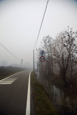 Kışın sisli bir günde çıplak ağaçlar ve tahta sütunlarla dar bir köy yolu.