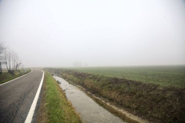 Kışın sisli bir günde, uzak bir yerde terk edilmiş bir evi olan bir kanal ve tarlalarla çevrili bir köy yolu.