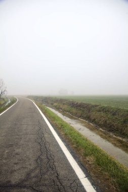Kışın sisli bir günde, uzak bir yerde terk edilmiş bir evi olan bir kanal ve tarlalarla çevrili bir köy yolu.