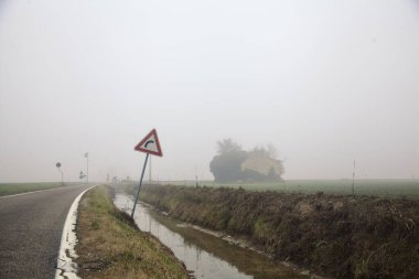 İtalya kırsalında sisli bir günde yolun kenarındaki bir tarlada terk edilmiş bir malikane ve sulama kanalı.