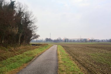 Asphalt trail bordered by a cultivated field and a forest next to a stream of water on a cloudy day in the italian countryside